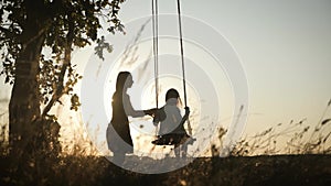 Silhouette of happy young mother and little daughter on a swing at sun light. Pretty girl sitting on a wooden swing and