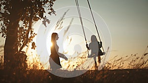 Silhouette of happy young mother and little daughter on a swing at sun light. Pretty girl sitting on a wooden swing and