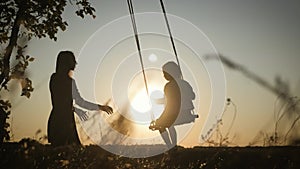 Silhouette of happy young mother and little daughter on a swing at sun light. Pretty girl sitting on a wooden swing and