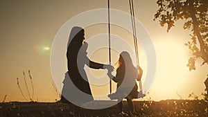 Silhouette of happy young mother and little daughter on a swing at sun light. Pretty girl sitting on a wooden swing and