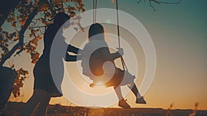 Silhouette of happy young mother and little daughter on a swing at sun light. Pretty girl sitting on a wooden swing and