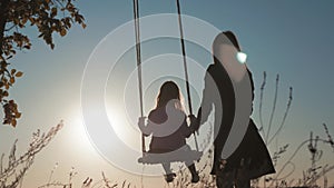 Silhouette of happy young mother and little daughter on a swing at sun light. Pretty girl sitting on a wooden swing and