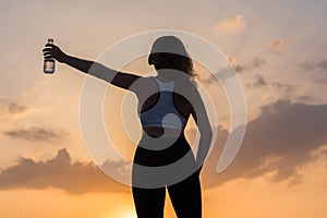 Silhouette of happy young woman in the sport clothes with bottle of clear mineral water on the sunset sky backgrounds. The concept