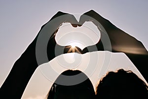 Silhouette, heart hands and couple at sky for sunset, love and care together on summer holiday with flare. Sign, closeup
