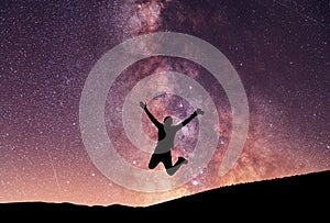 Silhouette of a hiker jumping above the hill in the starry night. Bright milky way galaxy behind him.