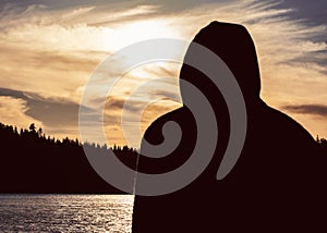 Silhouette of hooded man in golden sunset looking out over a lake.