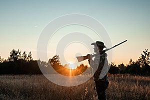 Silhouette of a hunter in a cowboy hat with a gun in his hands on a background of a beautiful sunset. The hunting period, the fall