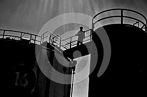 silhouette image of a refinery worker on the walkway between storage tanks.
