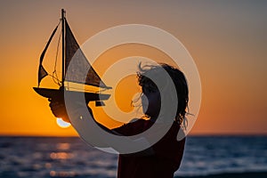 Silhouette of kid playing with toy seailing boat on sunset sea. Adventure and travel concept. Kid playing with toy