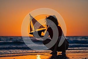 Silhouette of kid playing with toy seailing boat on sunset sea. Dream of summer. Kid play on the beach on a sunny day