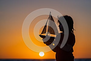 Silhouette of kid playing with toy seailing boat on sunset sea. Little boy playing with toy sailing boat, toy ship