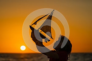 Silhouette of kid playing with toy seailing boat on sunset sea. Little kid boy enjoying summer vacation by the sea. A