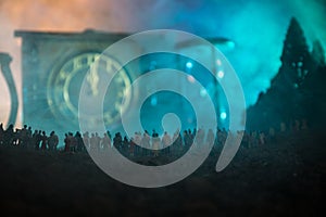 Silhouette of a large crowd of people in forest at night standing against a big arrow clock with toned light beams on foggy backgr