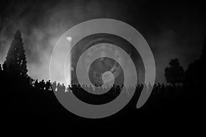 Silhouette of a large crowd of people in forest at night standing against a big arrow clock with toned light beams on foggy backgr