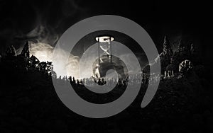 Silhouette of a large crowd of people in forest at night standing against a big hourglass with toned light beams on foggy backgrou