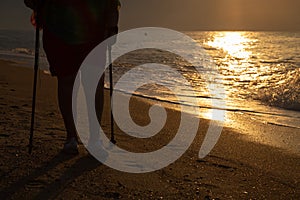 Silhouette of legs of an elderly woman doing nordic walking on the beach against sunrise