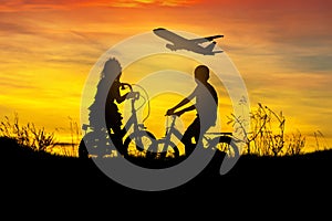 Silhouette little boy and little girl riding bike look to airplane flying on sunset
