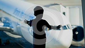 Silhouette of little boy looking on big airplane through big window in airport terminal