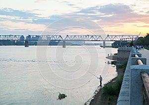 Silhouette of a lonely fisherman with a fishing rod on the river bank near the embankment in the evening against the background of