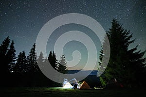 Silhouette of lonely hiker resting besides burning bonfire near illuminated tourist tents on camping site in dark