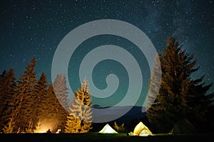 Silhouette of lonely hiker resting besides burning bonfire near illuminated tourist tents on camping site in dark