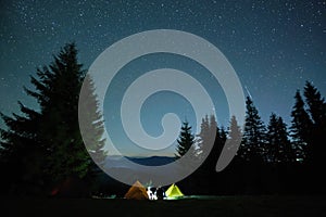 Silhouette of lonely hiker resting besides burning bonfire near illuminated tourist tents on camping site in dark