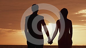 Silhouette of a loving couple at sunset. A man in love takes a woman by the hand and looks at the golden sunset on the