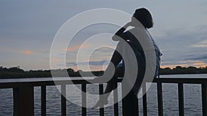 Silhouette of lyrical mood woman sitting on wooden rail of footbridge on lake at sunrise. Female person enjoying nature