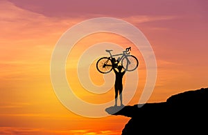 Silhouette the man in action lifting bicycle above his head stand on top of the mountain with sunset