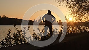 The silhouette of a man, against the backdrop of a river and a remote city. A person warms up before playing sports.