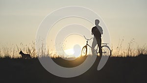 A silhouette of a man with a bicycle lifts his beloved dog to the top at sunset.