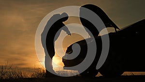 Silhouette man is checking engine bay of the broken down car while sunset.