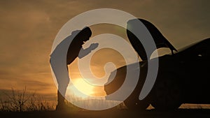 Silhouette man is checking engine bay of the broken down car while sunset.