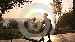 Silhouette of a man doing arm exercises and squats outdoors using resistance band outdoors in nature. Triceps and legs
