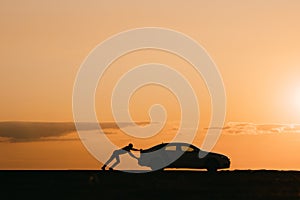 Silhouette of man driver pushing his car along on empty road after breakdown at sunset