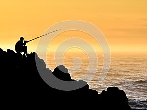 Silhouette of man fishing whilst sitting on coastal rocks.