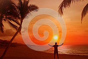 Silhouette of a man with his arms outstretched. Behind him is a beautiful sunset over the sea and the beach with palms