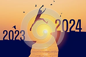 Silhouette man jumping between cliff with number 2023 to 2024 and birds flying at tropical sunset beach. Freedom challenge and