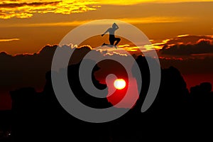 Silhouette of man jumping over cliff  above the clouds and the sun on sunset background