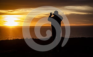 Silhouette of a man looking out over the ocean taking a cell phone picture at a sunset