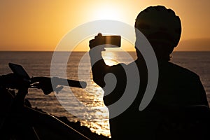 Silhouette of man looking at sunset taking a photography with mobile phone, wearing sport helmet after cycling along the sea