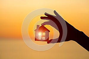 Silhouette of man`s hand holding small wooden house against sunset or sunrise light