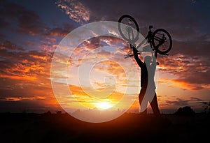 Silhouette the man stand in action lifting bicycle above his head on the meadow with sunset.