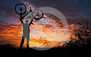 Silhouette the man stand in action lifting bicycle above his head on the meadow