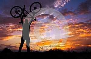 Silhouette the man stand in action lifting bicycle above his head on the meadow with sunset