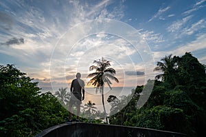 Silhouette of man standing alone on the hill with a coconut palm trees and enjoys sea on a horizon in a sunset. Vacation