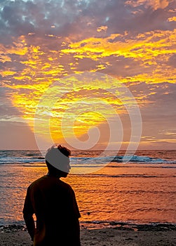 Silhouette of a man standing on the beach against the background of the sunset sky emitting a golden triangular light at dusk.