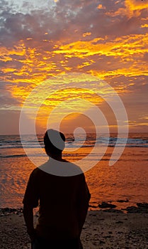 Silhouette of a man standing on the beach looking at the sunset which emits a golden triangular light at dusk.