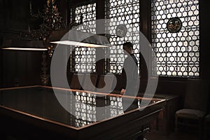 Silhouette of man standing by a grand table in an old house. Leaded windows letting in light from behind him.