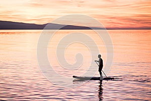 Silhouette of man at sunset standing on paddle board. Summer beach leisure activity.